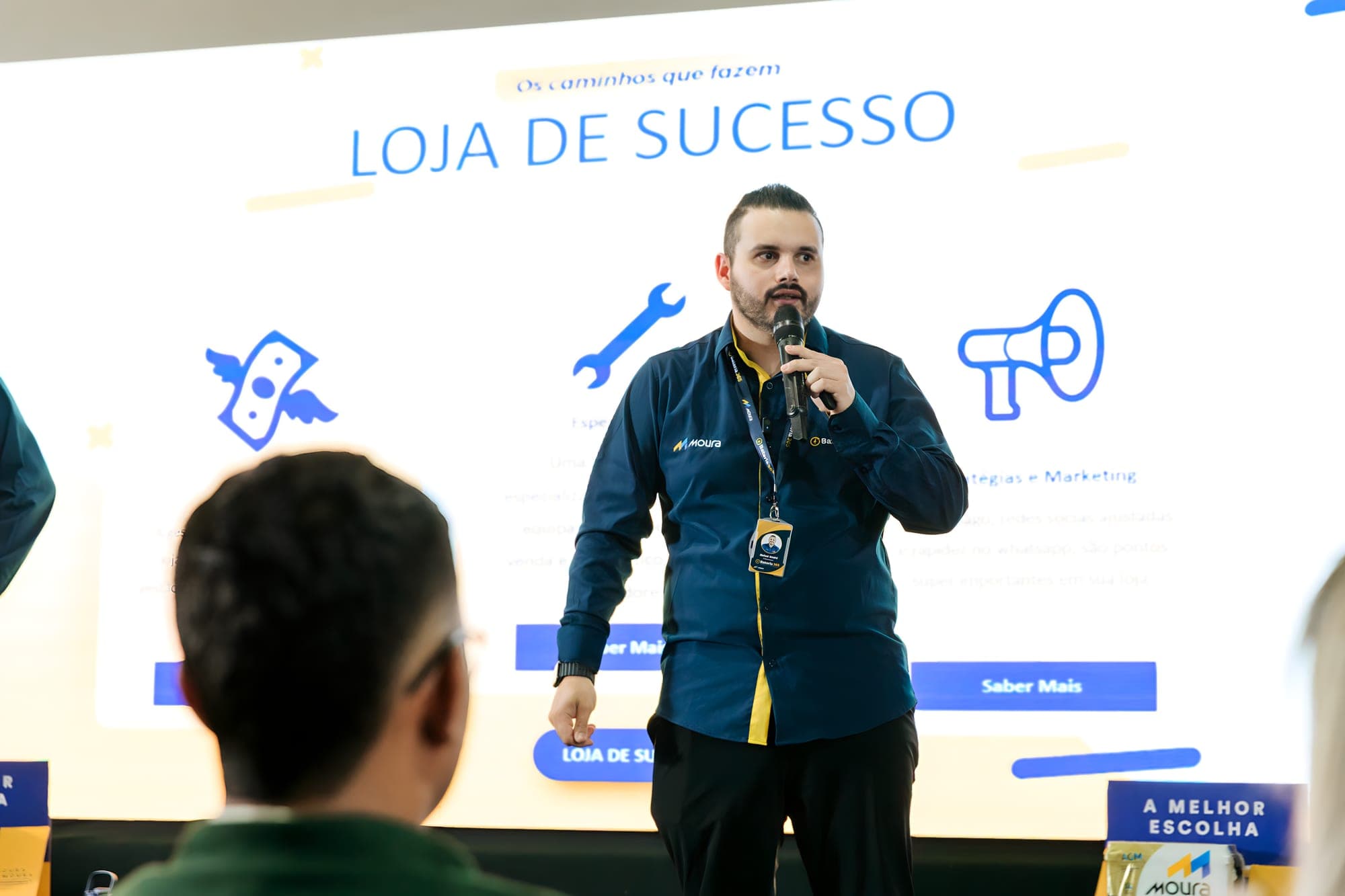 Rafael palestrando sobre loja de sucesso em evento da Baterias Moura, com o telão ao fundo.
